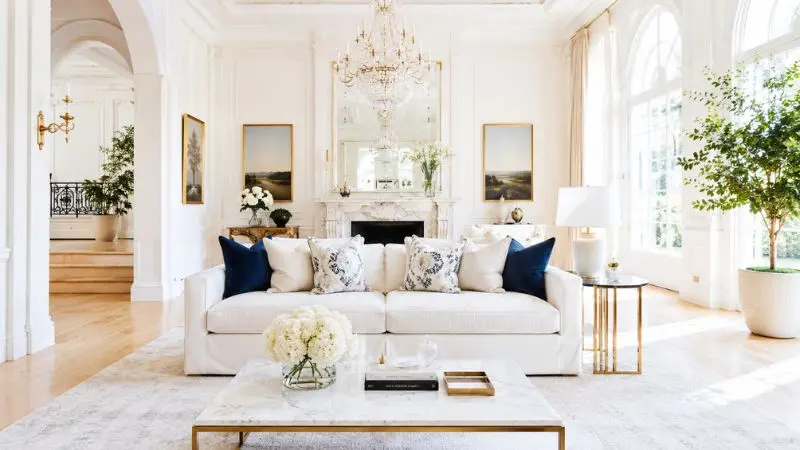 A bright and spacious luxury classic living room with white wall moldings, a crystal chandelier, and a marble fireplace, featuring a stylish white fabric slipcover sofa decorated with elegant deep blue and patterned throw pillows in the center.
