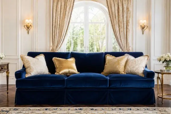 A luxury classic well-lit living room featuring a premium rich navy blue velvet slipcover sofa decorated with elegant gold and patterned silk cushions on a dark herringbone wood floor.