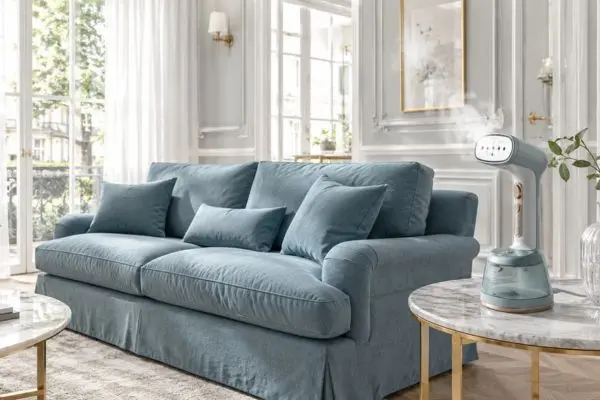 A bright luxury classic living room featuring a premium soft teal blue fabric slipcover sofa with a modern handheld garment steamer releasing steam on a marble side table.