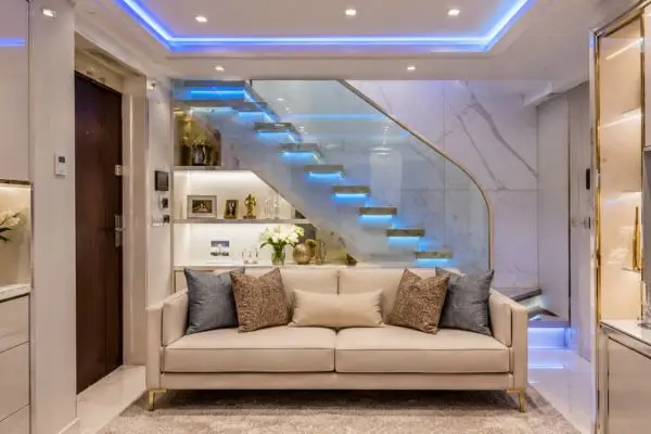 Compact cream sofa under a glass staircase with blue LED lights in a small room.