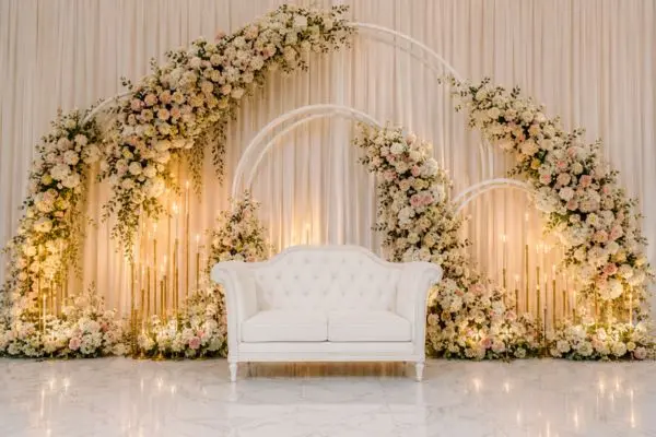 Elegant white tufted two seater furniture on a luxury stage background with blush rose arches and gold candles.